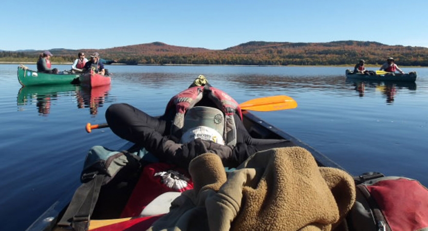 From the back of a canoe, you can see the person in front leaning back in relaxing fashion. There are additional canoes floating on calm water in the background.